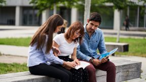 Three students sit on a wall on campus and look at information material.
