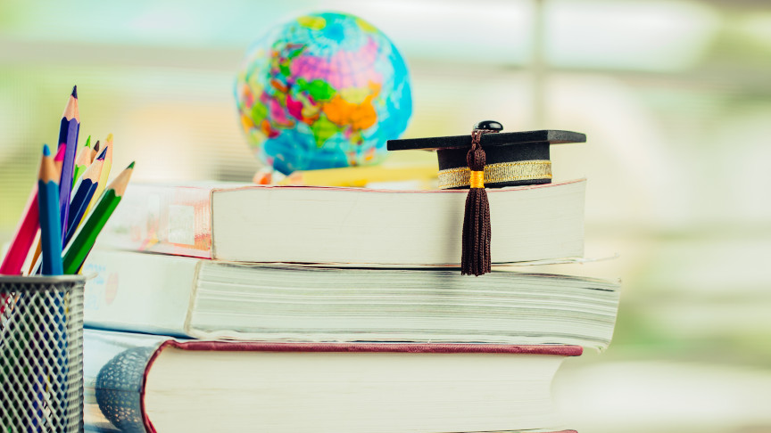 Schwarzer Doktorhut auf einem Stapel Lehrbücher neben einer Bleistift-Box und einem Globus im Hintergrund